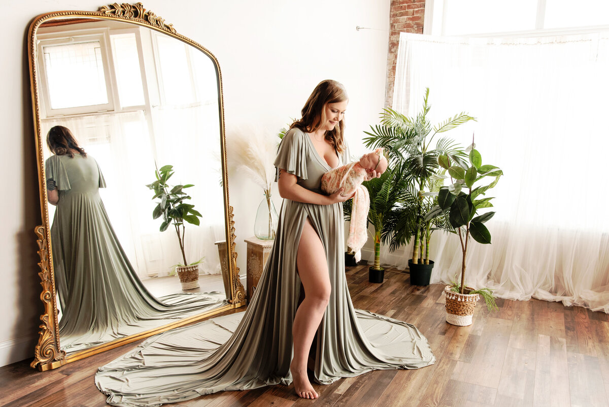 st-louis-newborn-photographer-mom-in-green-gown-holding-baby-girl-in-front-of-large-mirror-and-plants