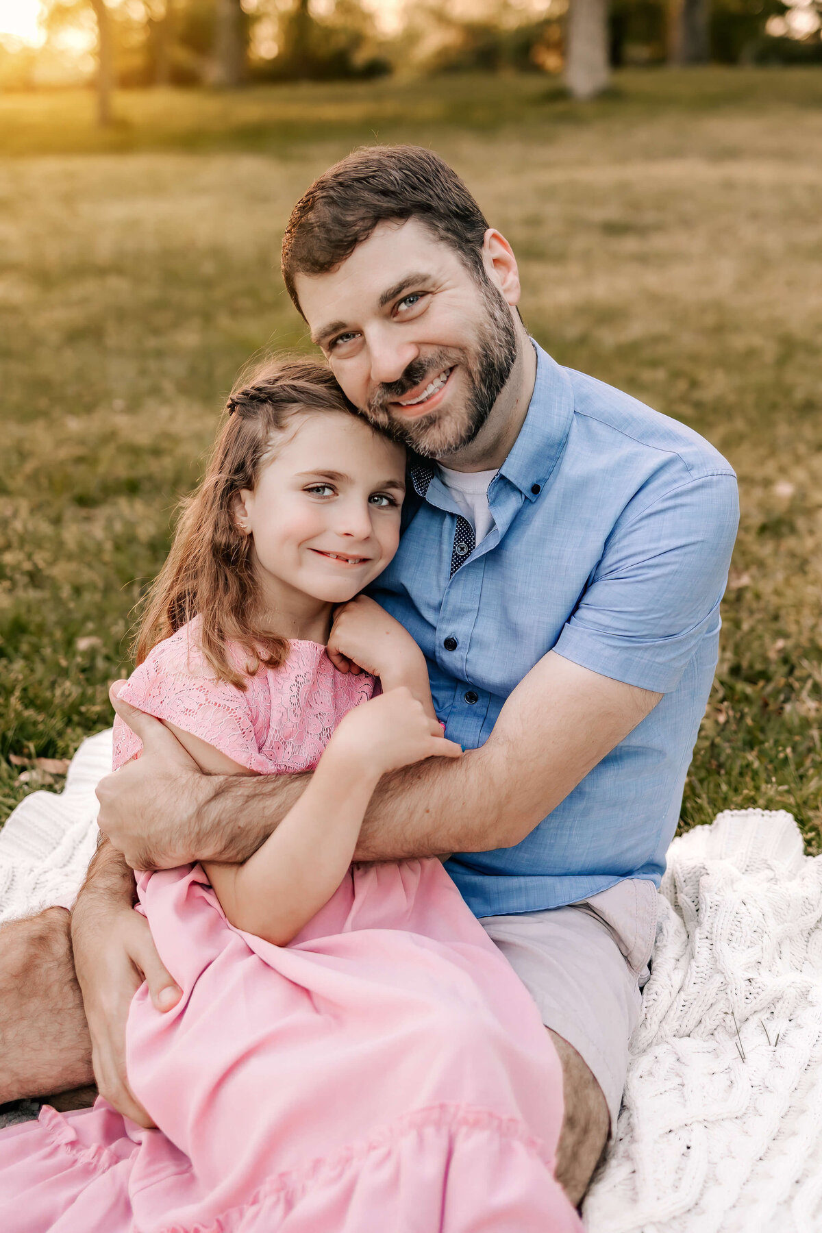 An adorable girl is sitting in her dads lap as they wrap their arms around each other and snuggle in for their family photography session with Foppiano Photography.