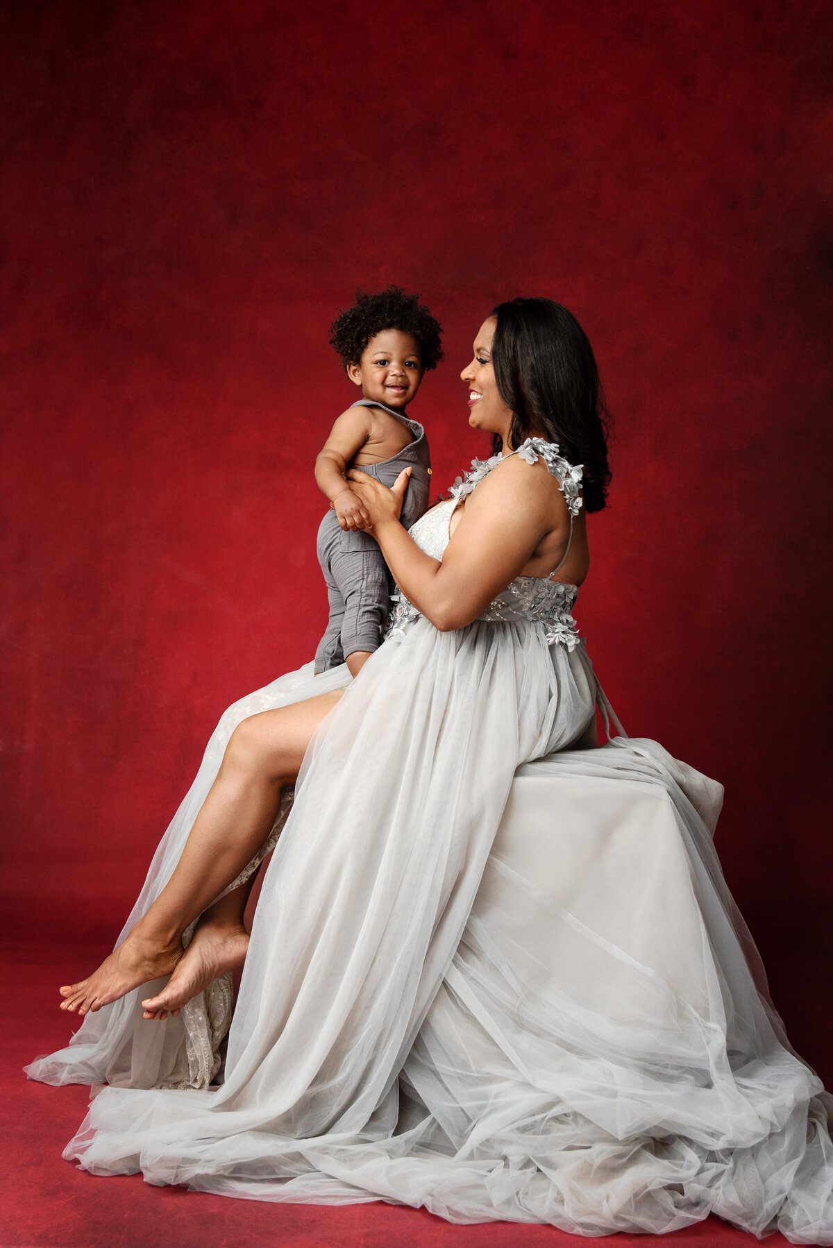 st-louis-family-photographer-mom-in-gray-tulle-gown-sitting-with-holding-todder-boy-on-red-backdrop