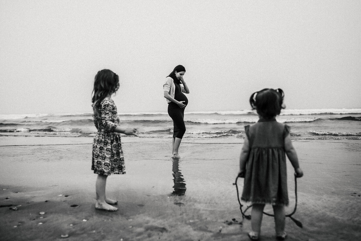 Pregnant mom holds belly on beach while two young girls look in her direction
