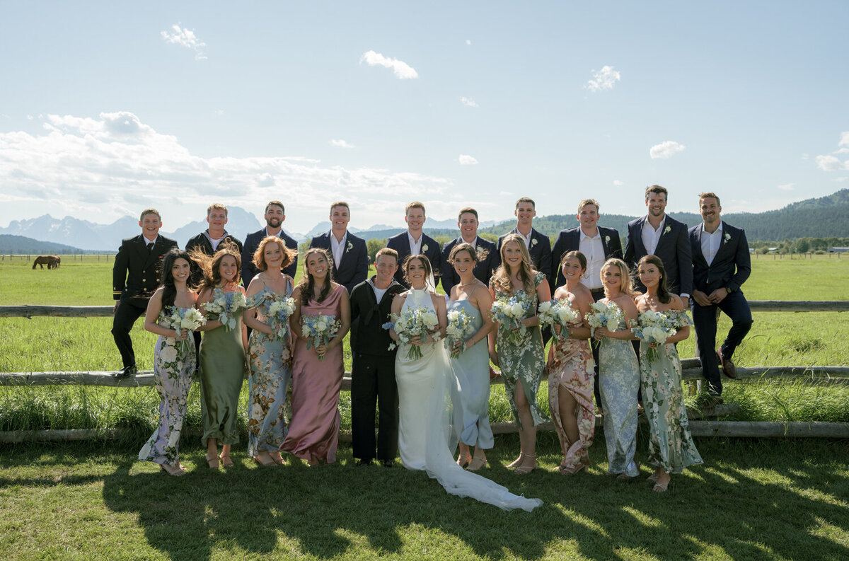 Bridal party posing along a rustic wooden fence with the majestic Teton mountain range in the background, captured during a sunlit summer wedding in Jackson, Wyoming.