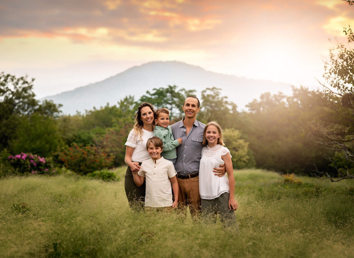 A mom, dad and their children standing in tall green grass with flowers in the background and smiling during their Asheville Family Photographer portrait session
