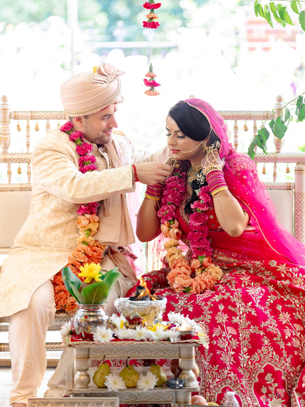 A couple in traditional Indian attire participates in a wedding ceremony. The groom is placing a necklace on the bride. Both are adorned with floral garlands. They sit beside a decorated altar with a sacred fire and flowers.