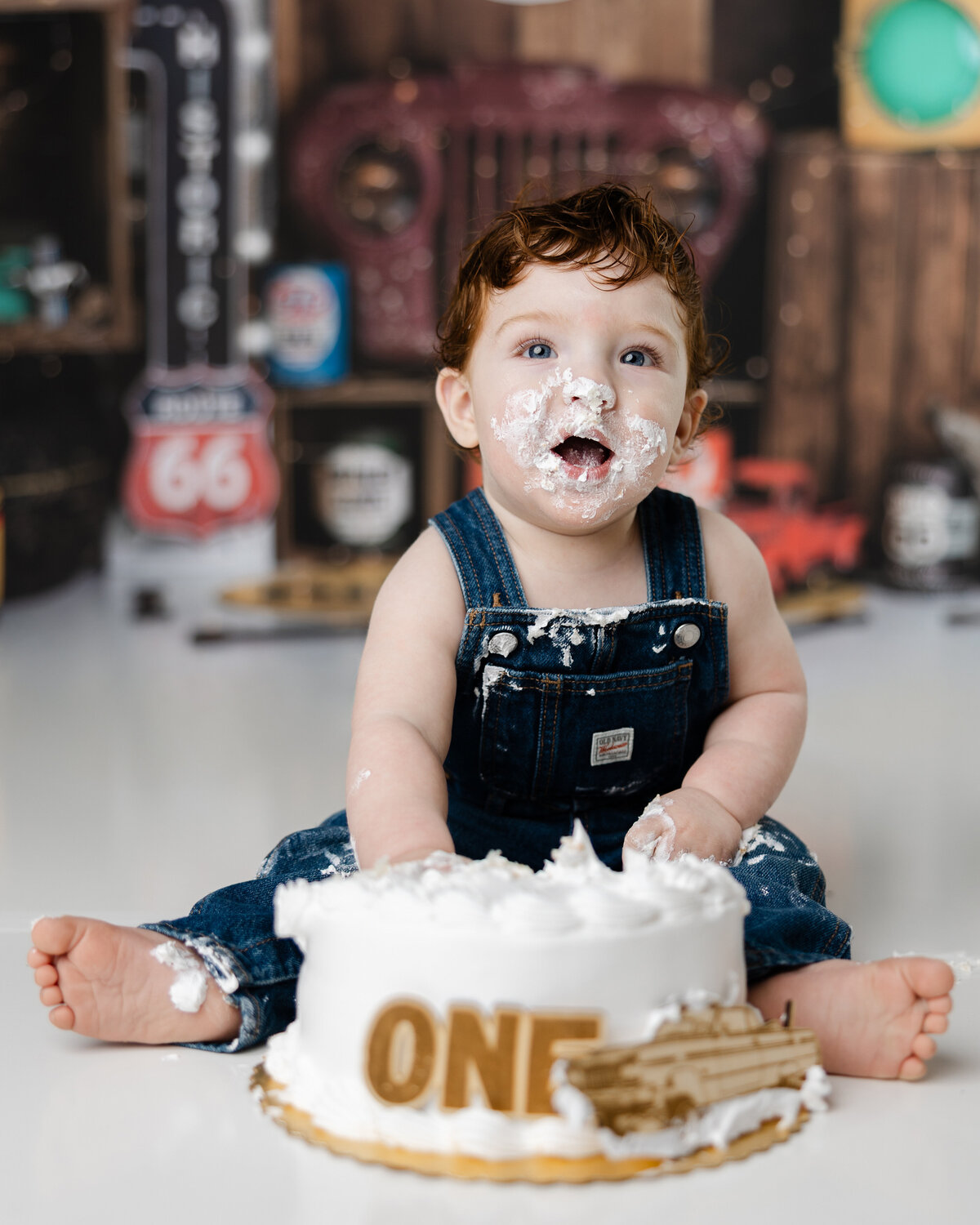 Little boy celebrating his first birthday with a cake smash, face covered in frosting and set against a retro garage theme.
Photograph by Yucaipa cake smash photographer Kaitlyn Dawn Photography.