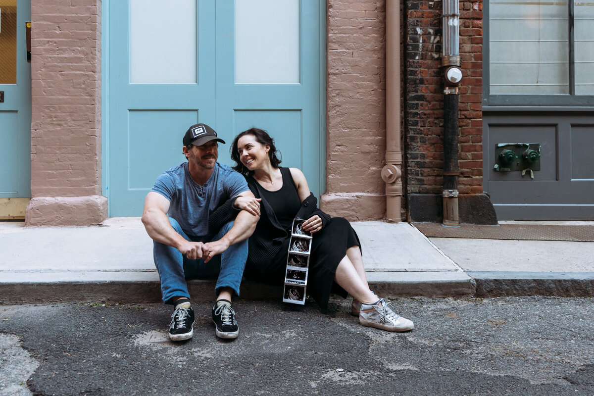Maternity Photographer, a pregnant woman and man sit on the city curb laughing together
