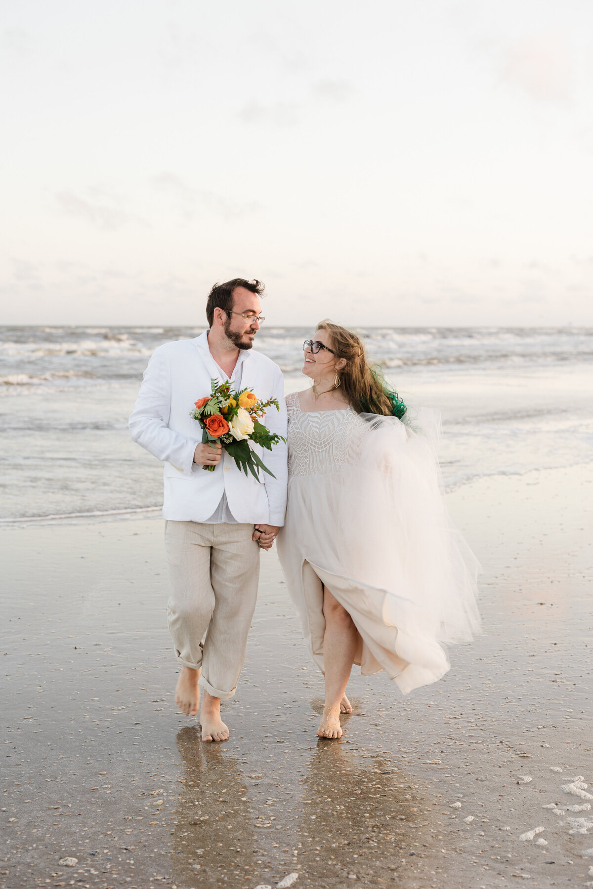 Portrait of a bride and groom walking along the beach and looking longingly at each other after their wedding ceremony on the beach in Crystal Beach, Texas. The bride is on the right and is wearing a detailed flowing white dress with green highlights in her hair. The groom is on the left and is wearing a white jacket and khaki pants while holding the bride's bouquet. They both are wearing glasses and are backed by the ocean.