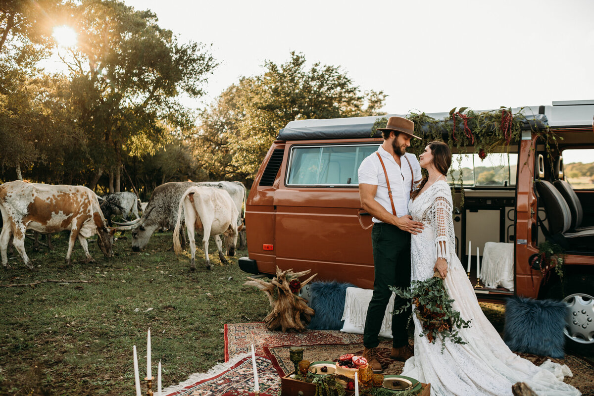 Couples Photography, Man in suspenders and a brown hat holds woman in a wedding dress, in front of a vintage VW bus with longhorns in the background.