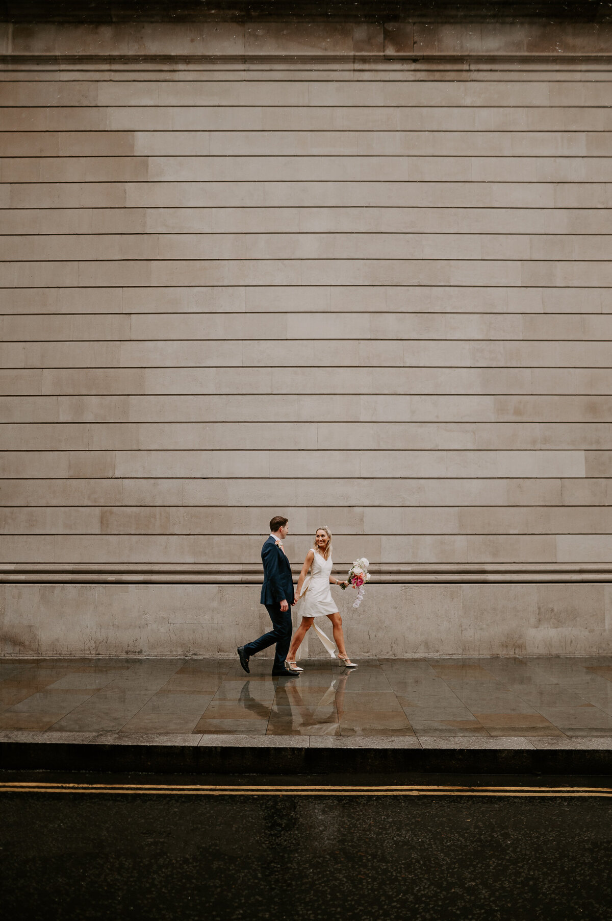 A wedding couple walk through Bank in London near the Ned, the Bride is wearing a short wedding dress and carrying a pink bouquet.