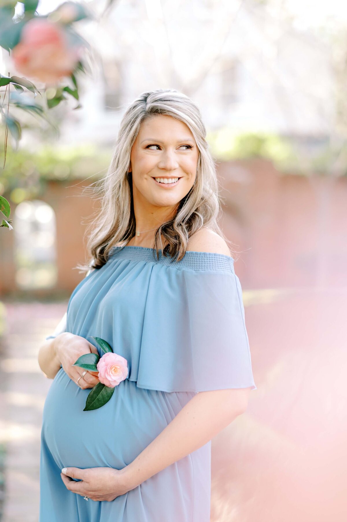 A beautiful blonde woman in a blue dress smiles off to the distance as she hugs her pregnant belly under camellia tree blooms.  Captured by Charlotte Maternity Photographer Melissa Mayrie Photography.
