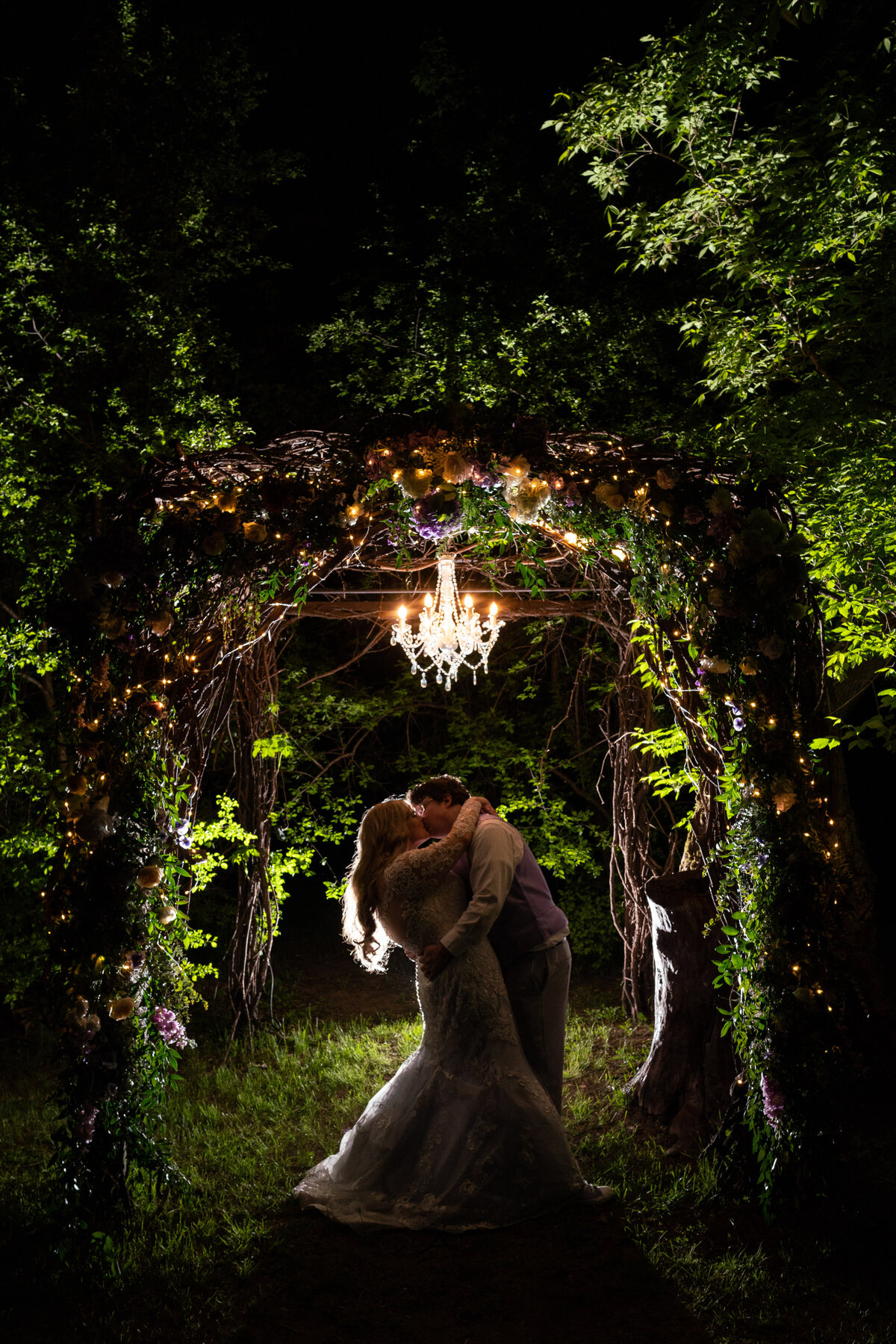 Bride and groom kiss under lights in a garden at night.