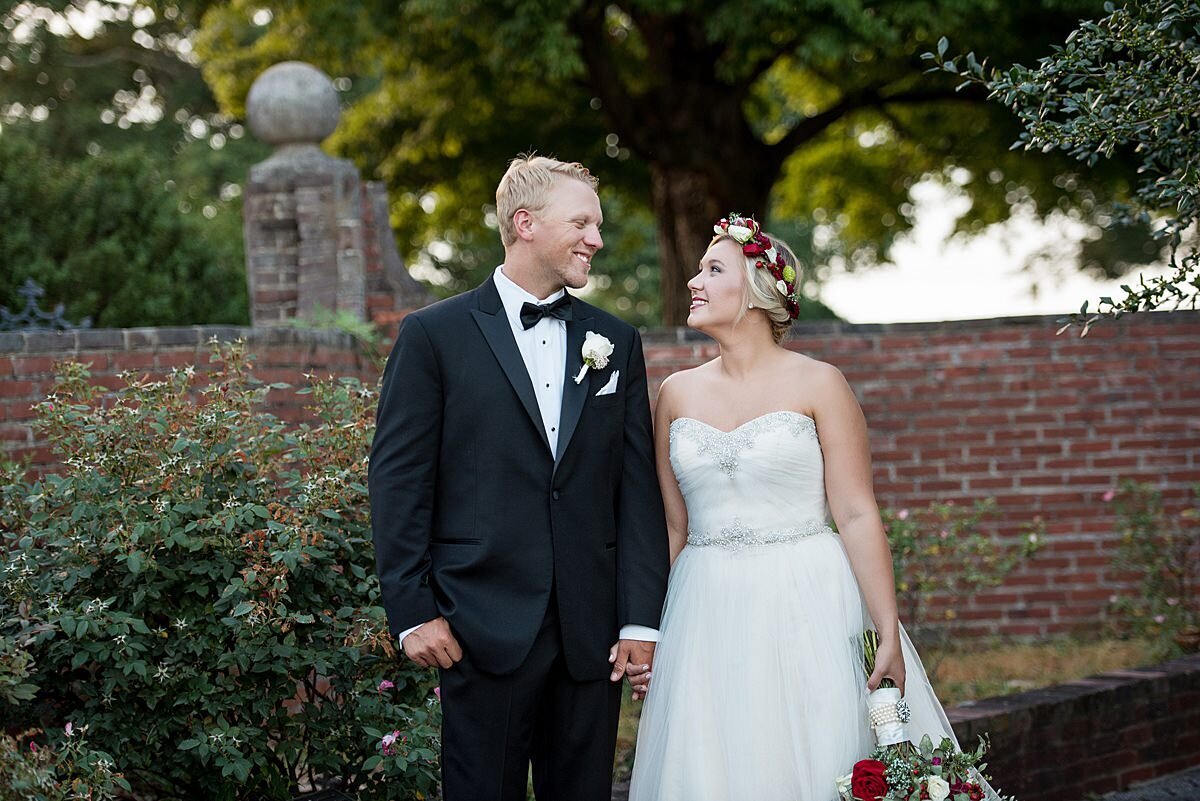 The bride and groom hold hands in front of a low brick wall. The groom is wearing a black tuxedo with a white shirt and black tie. The bride is wearing a strapless wedding dress with a sweetheart neckline and a rhinestone belt. Her blush, pink and red bouquet is being held down at her side.