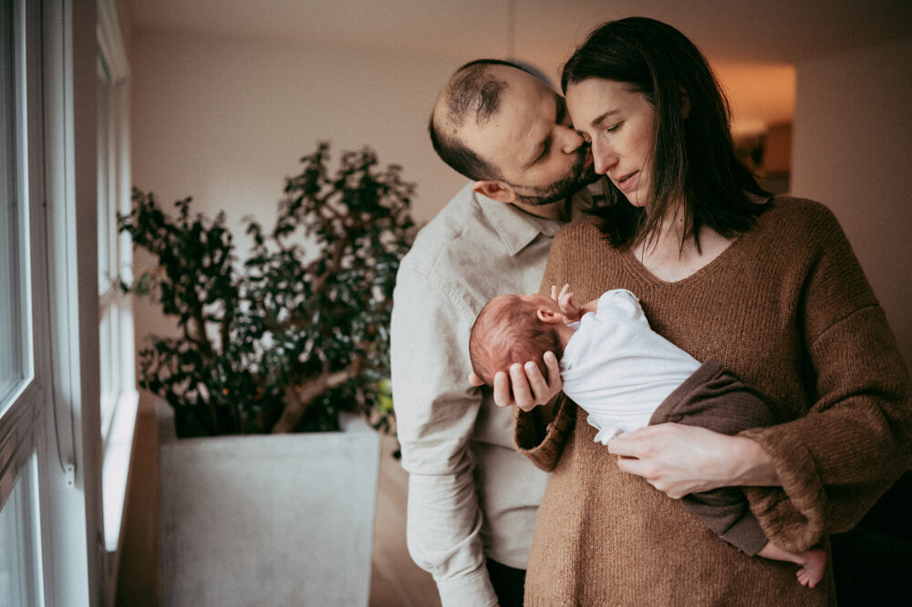 Tender family moment—father kissing mother’s forehead while she holds newborn, captured during a calm, emotional in-home newborn session in Portland