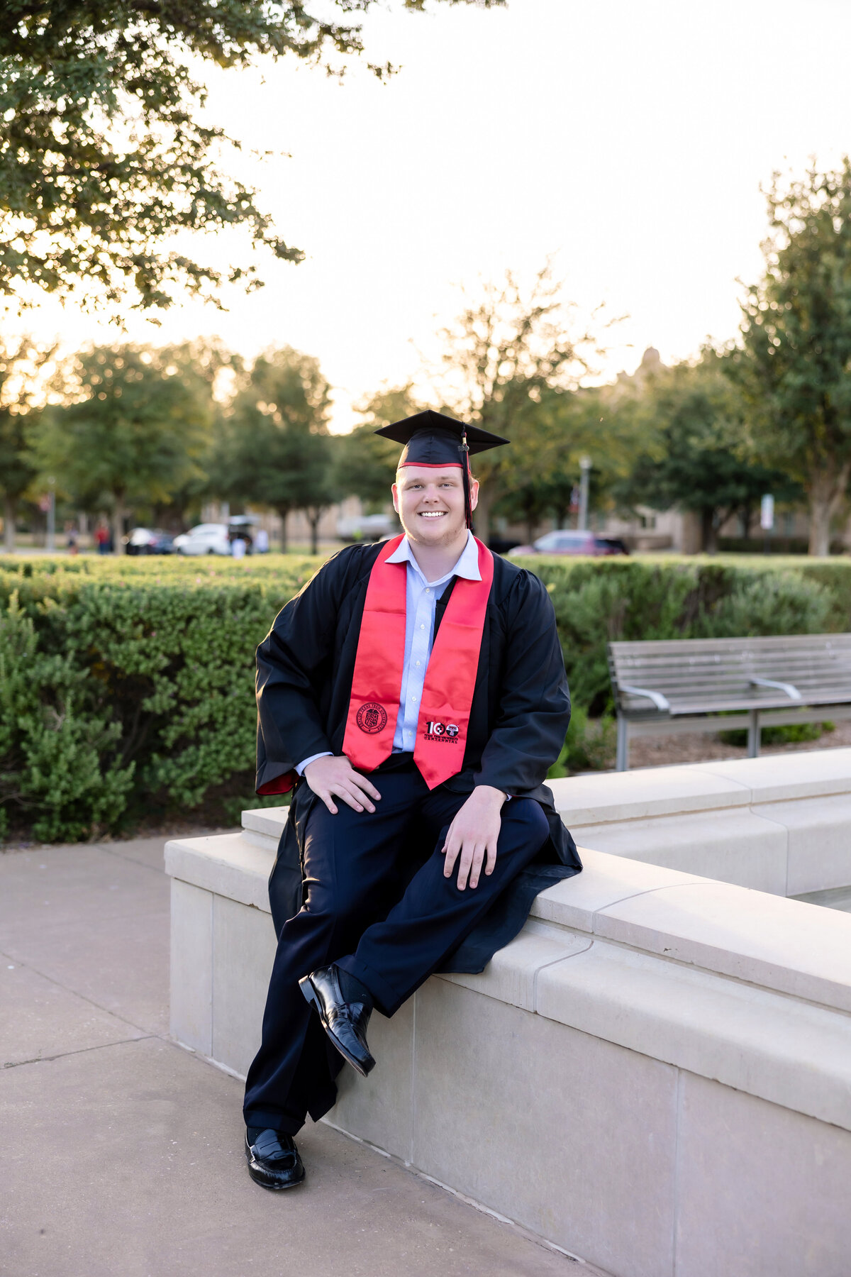 College male sitting on the TTU fountain and wearing a graduation gown with red stole and cap with tassel while smiling and looking at the camera