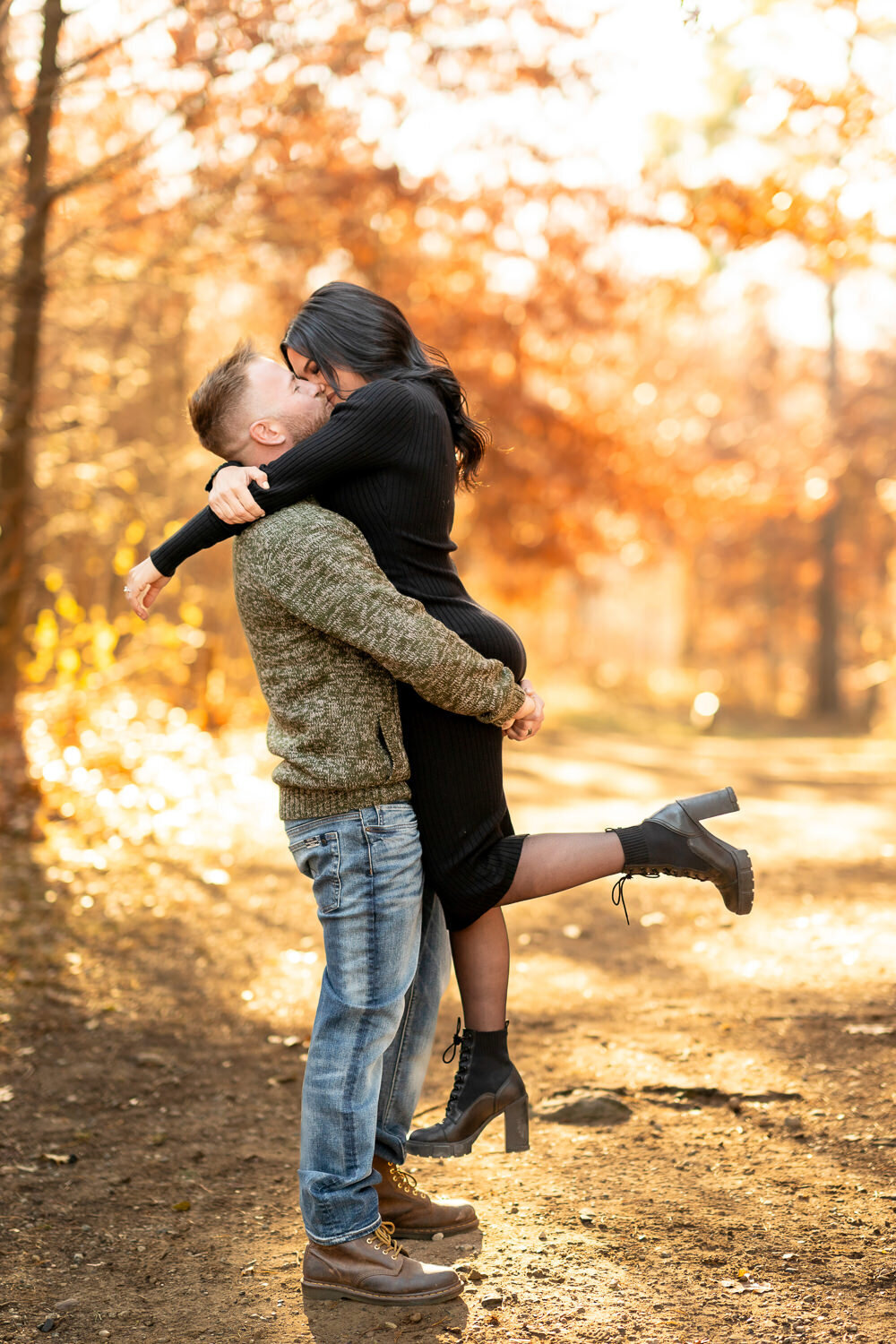 Man lifts and kisses woman in black dress with fall colors in the background.