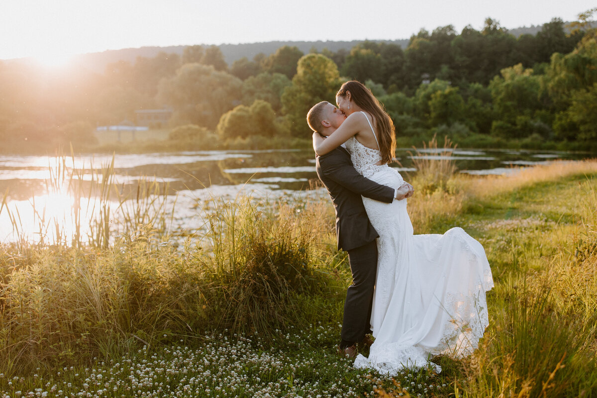 groom holding up his bride in a grassy area by water