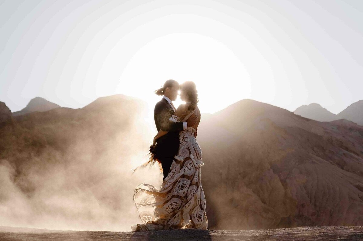 A bride and groom share a moment, backlit by the sun with dust swirling around, and mountain peaks behind them.