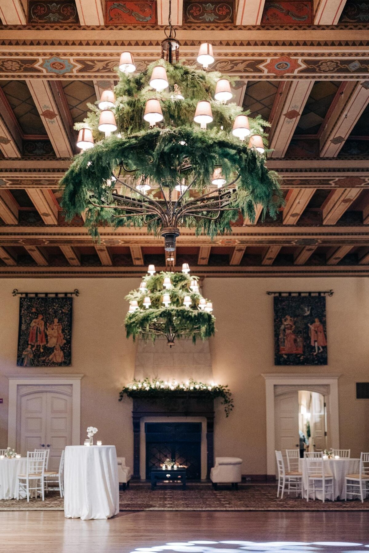 An elegant chandelier adorned with greenery hangs in a grand hall with a fireplace and tapestries.