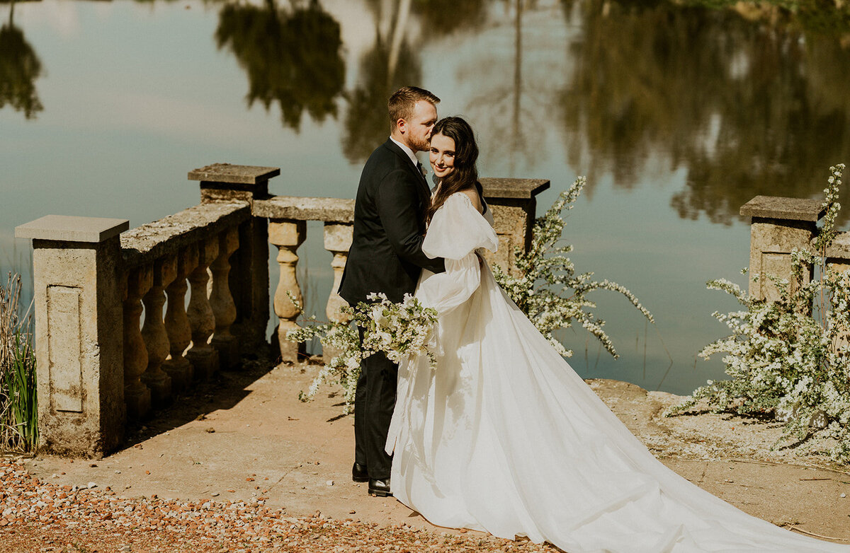 the-groom-is-kissing-bride-on-the-cheek-while-she-looks-into-the-camera-near-the-lake