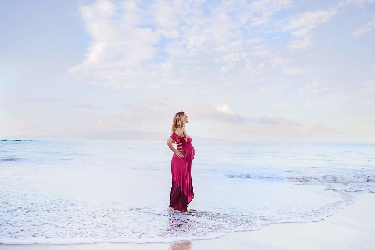 Woman poses with her hand on her hips and holds her pregnant belly for her Wailea maternity photoshoot