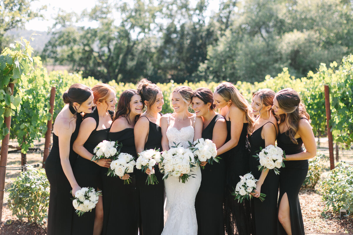 bride with her bridesmaids dressed in black cocktail dresses smiling together in sonoma vineyard.
