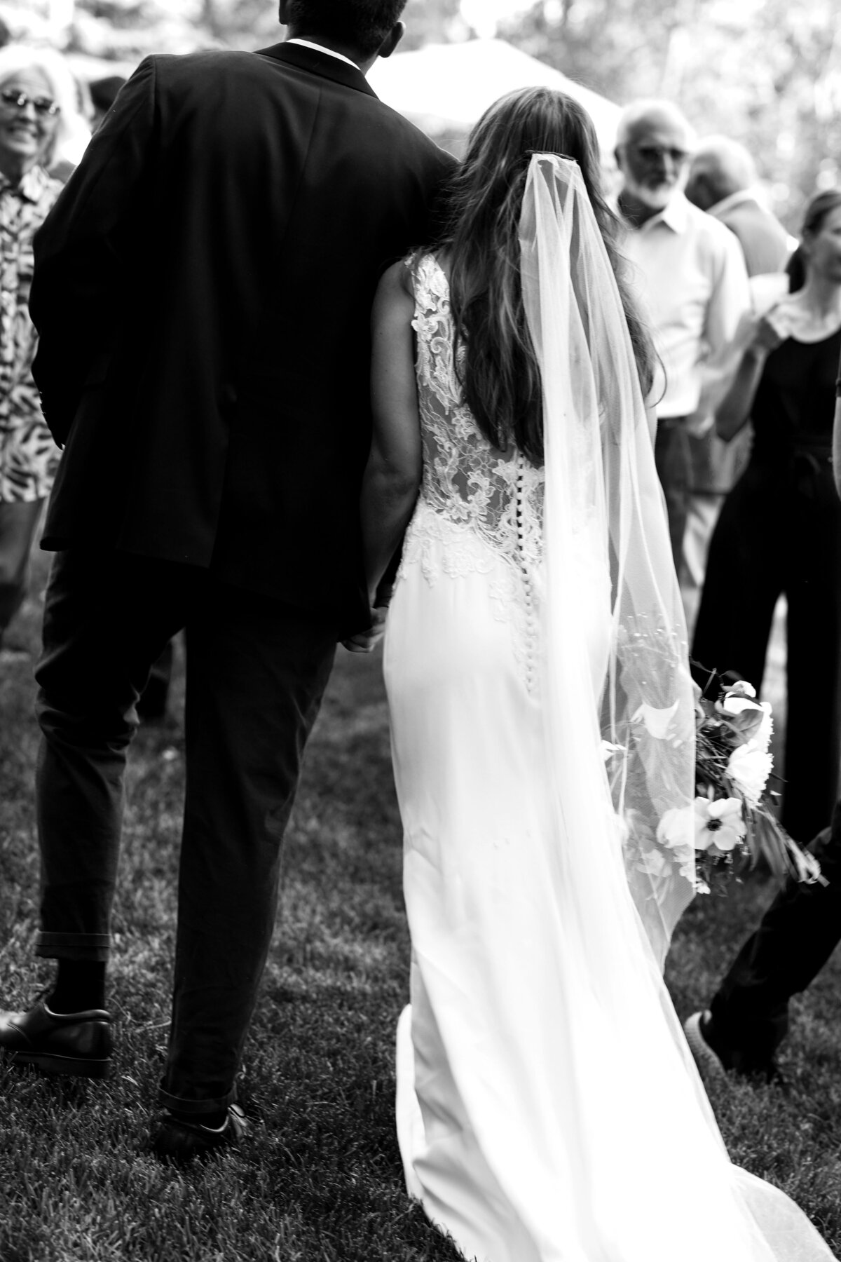 A black and white portrait of a bride and groom walking towards their guests, with a view of the brides white dress from the back with lace and buttons.