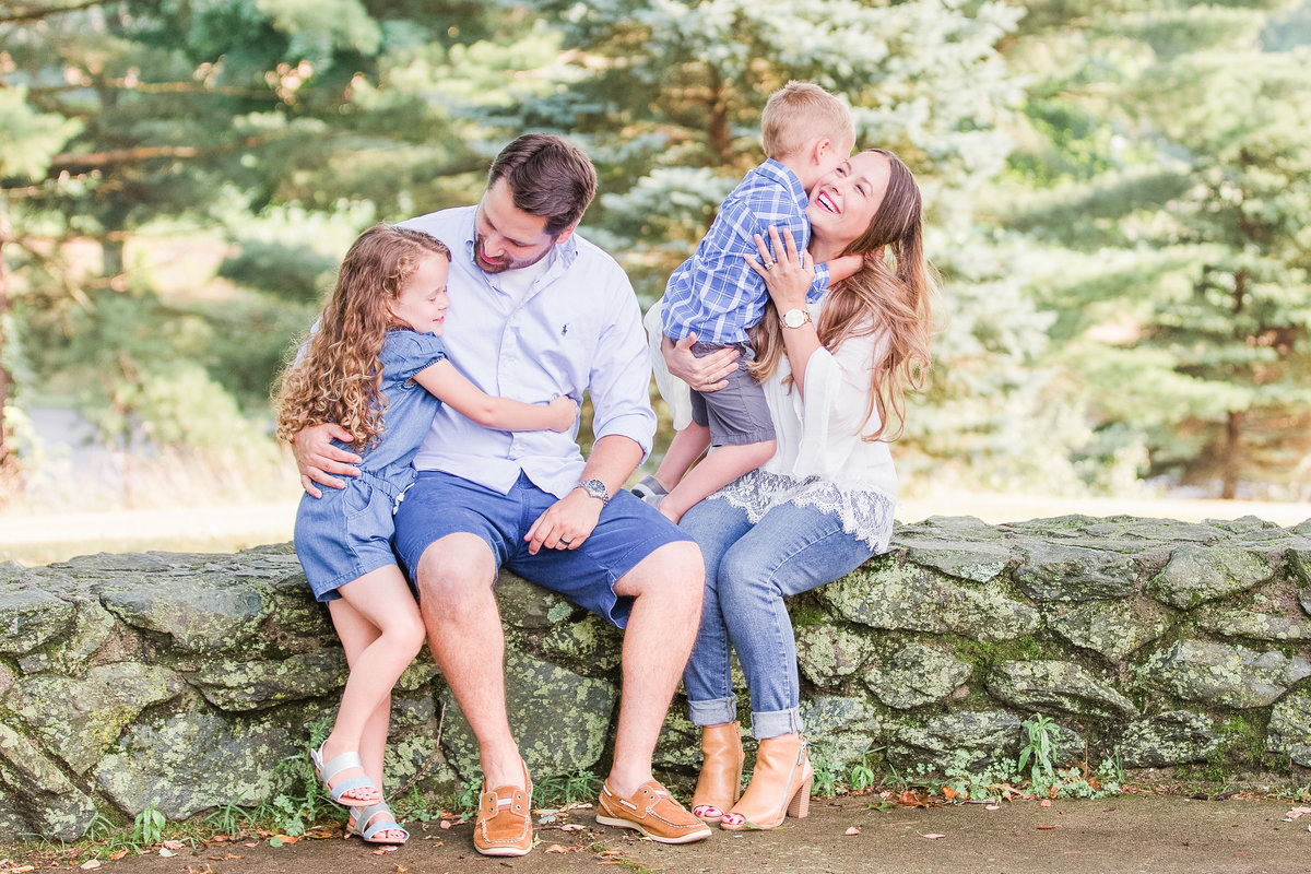 Mom and dad embrace their two young children while sitting on a stone wall outside