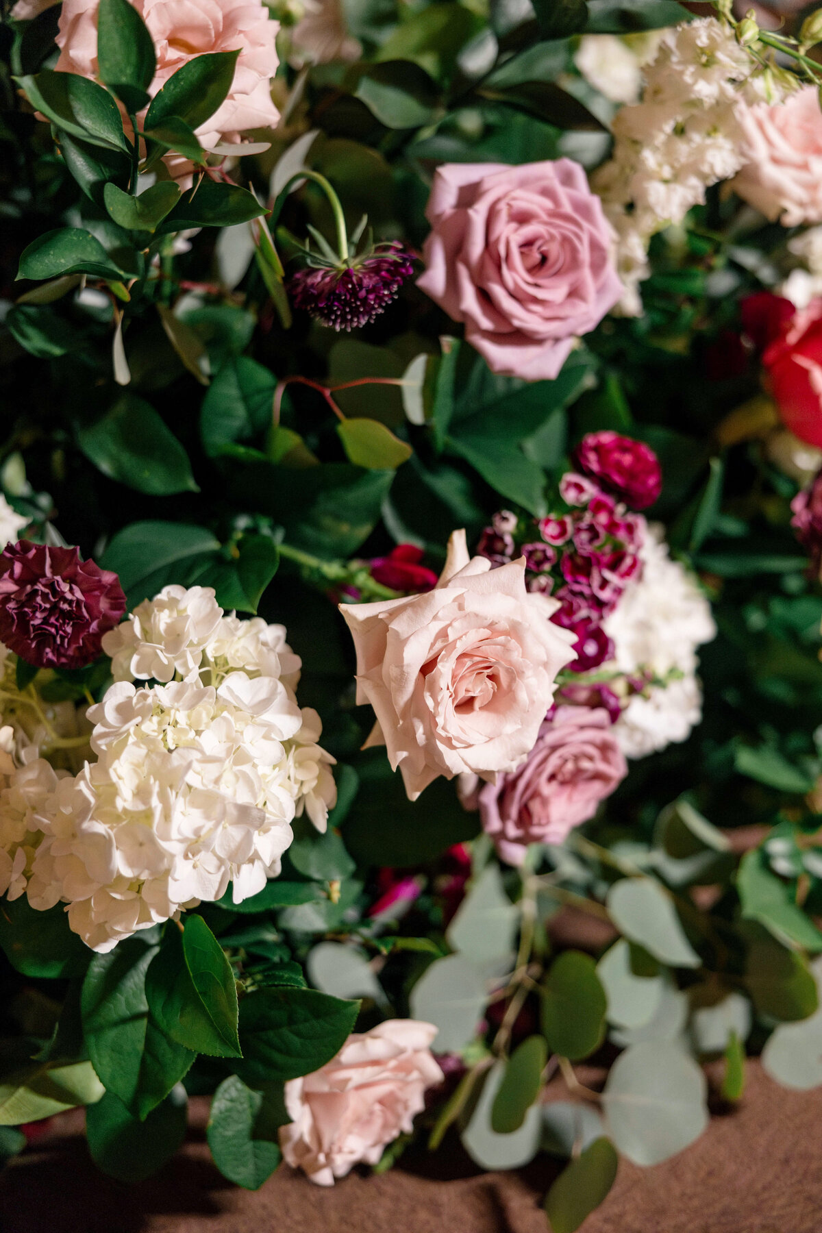 A lush floral arrangement featuring pink roses, white hydrangeas, burgundy flowers, and abundant green leaves. The contrasting colors and textures create a rich and elegant display.