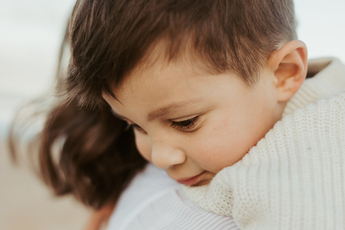 A close up of a little boy in a cream knitted jumper over his mum's shoulder.