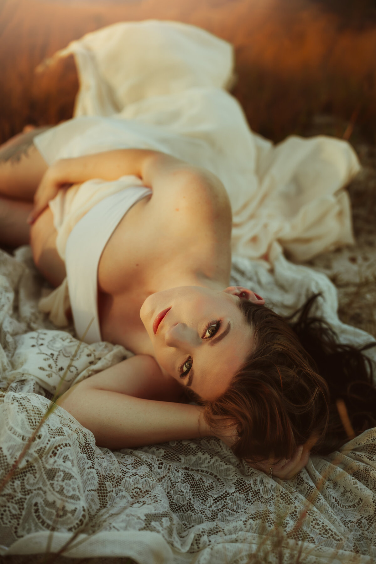 timeless maternity portrait of a women laying on a vintage lace blanket in tall grass on the bacj as sunset hugging her baby bump looking at the camera at sunset with glowy golden hour light