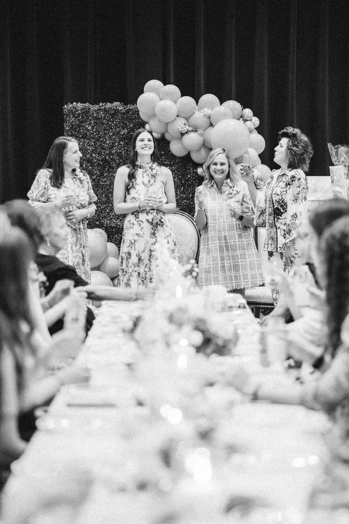 Black and white image captured by an event photographer in Fayetteville, NC, shows four women standing and smiling at the head of a long table. The table is elegantly set for a meal, with guests seated and focused on the women. A decorative balloon arrangement adorns the background.