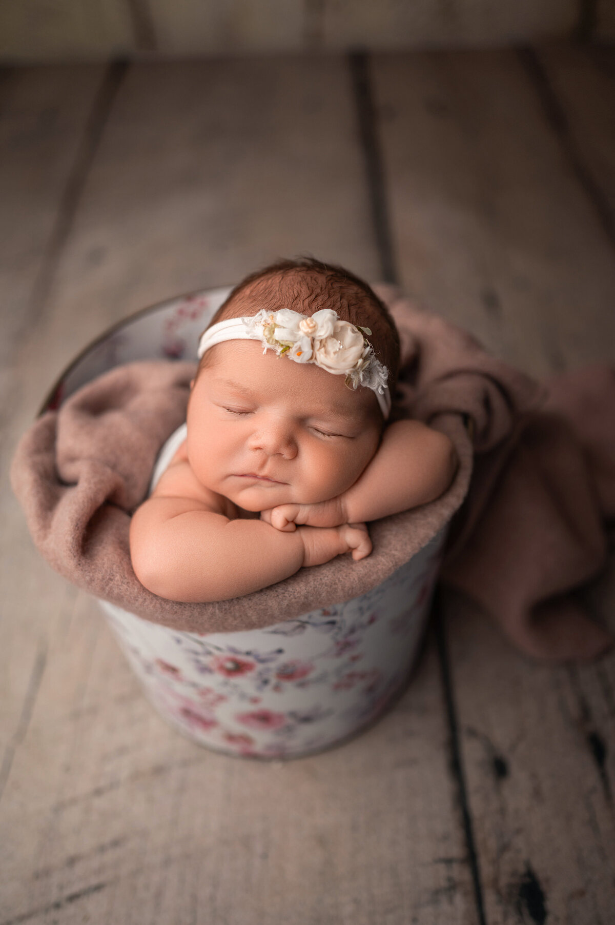 Portrait of a baby girl asleep in a  metal bucket with floral print. Baby is wearing a floral headband and wrapped in a soft, neutral blanket.