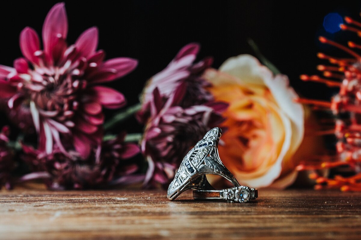 Sitting on a wood table in front of purple mums and orange roses sits an antique art deco diamond engagement ring and a white gold wedding band with a solitaire diamond.