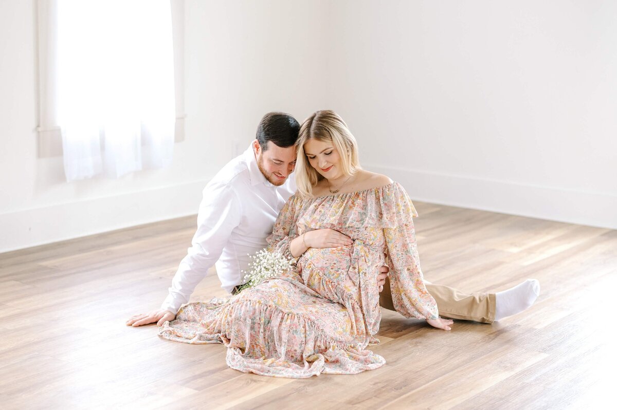 A husband and wife snuggle on the floor of a natural light studio while looking down at her pregnant belly in  Charlotte, NC.