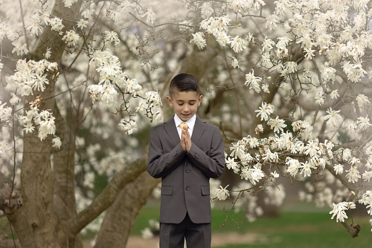 A young boy prays in a grey suit while standing under a while flowering tree