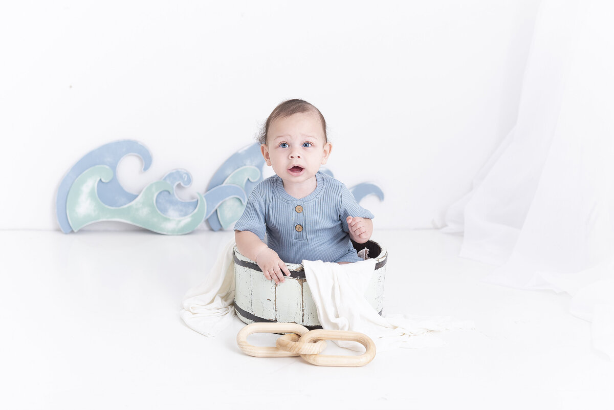 A toddler boy in a blue onesie sits in a bucket in a nautical themed studio of a childrens photographer Atlanta