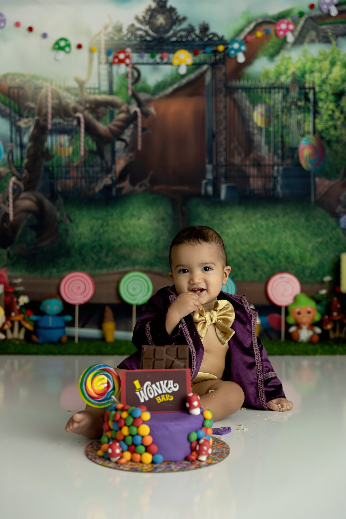 A toddler boy in a gold bowtie and purple jacket sits for a Wonka themed first birthday session with a  New Jersey Milestone Photographer