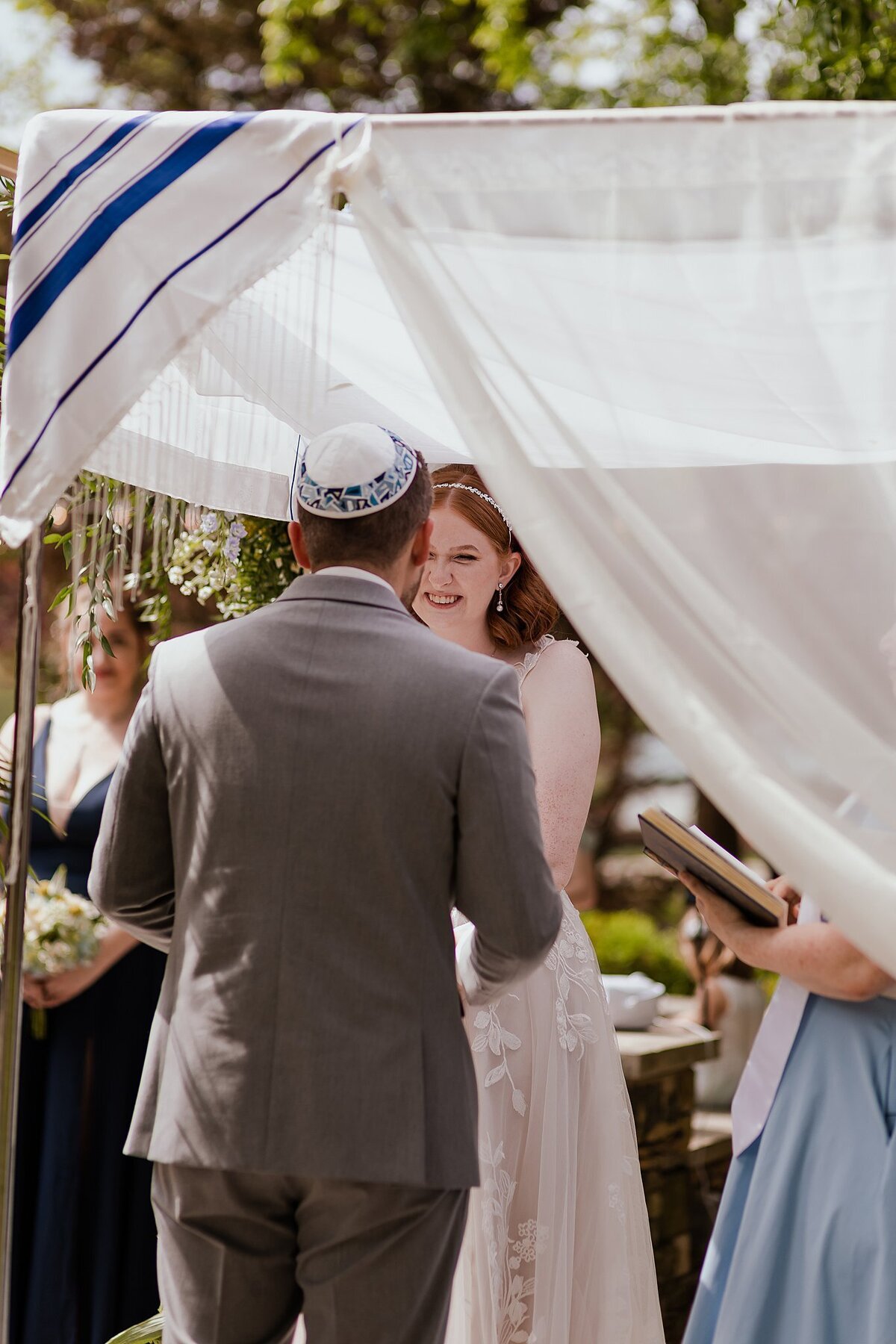 Red haired bride wearing a white lace wedding gown smiles at the groom wearing a blue and white kippot and light gray suit while standing under the chuppah with their officiant in a light blue dress.