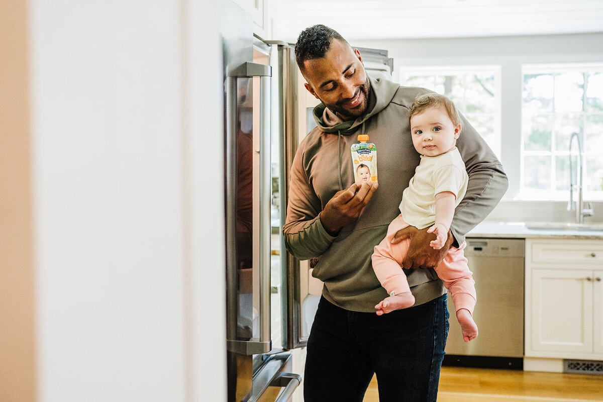 dad holds baby in arms while getting yogurt from fridge