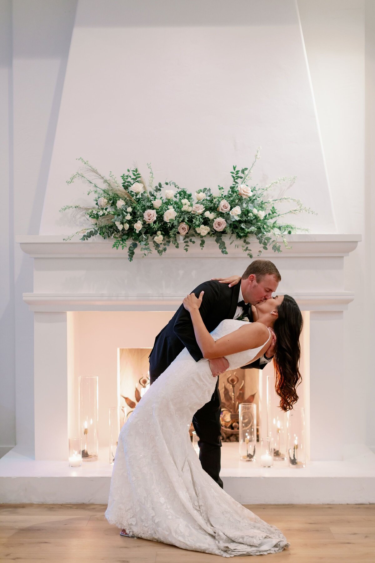 The bride and groom standing in front of a fireplace with a large white floral piece above.