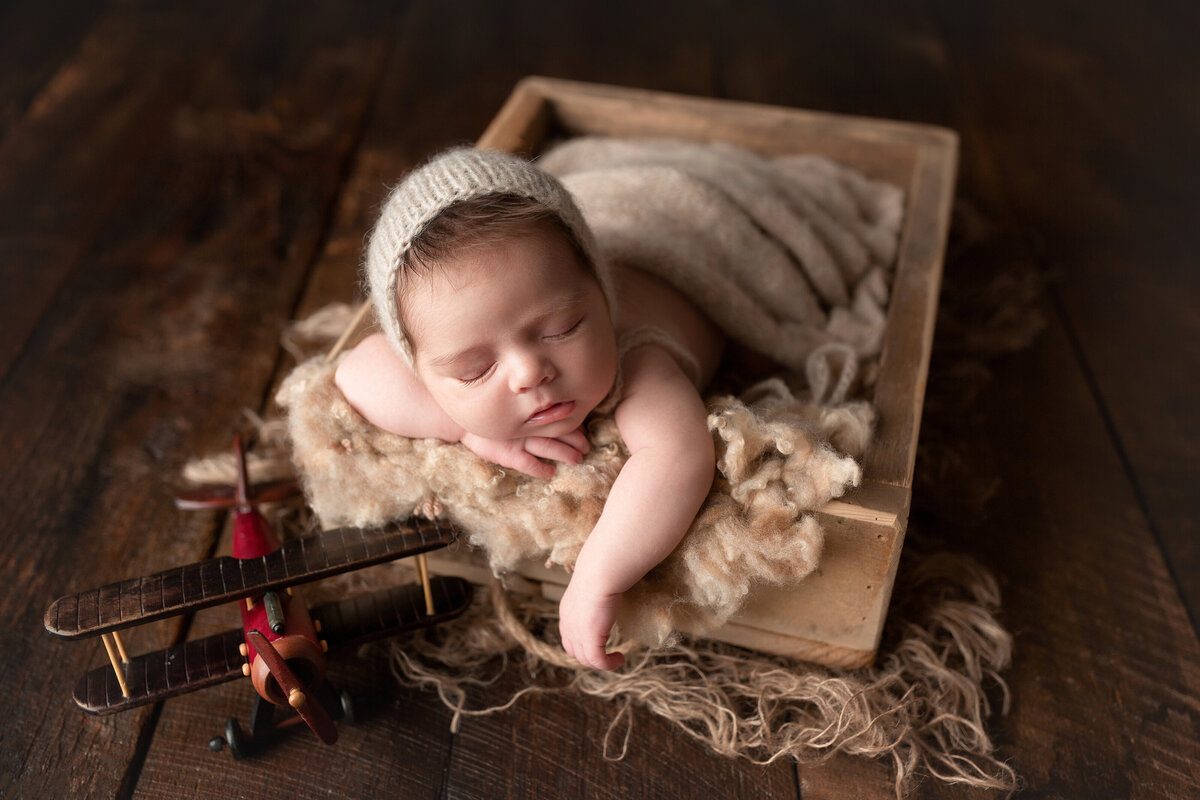 Baby boy sleeping in a small crate. Baby's right hand is resting under his chin and the left hand is draped over the edge of the crate. Baby is resting his cheek on his right arm. There is a vintage toy play beside the crate.