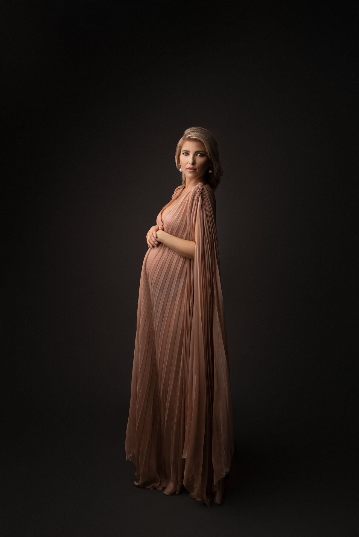 Woman poses for fine art maternity photos with Philadelphia's best maternity photographer Katie Marshall. Woman in a long dusty rose maternity photoshoot gown with floor-length caped sleeves stands side profile to the camera. Both of her hands are resting atop of her bump. She is looking over her shoulder intently at the camera. Light envelops the left side her body is facing, her back side shadowed creating depth to the image.