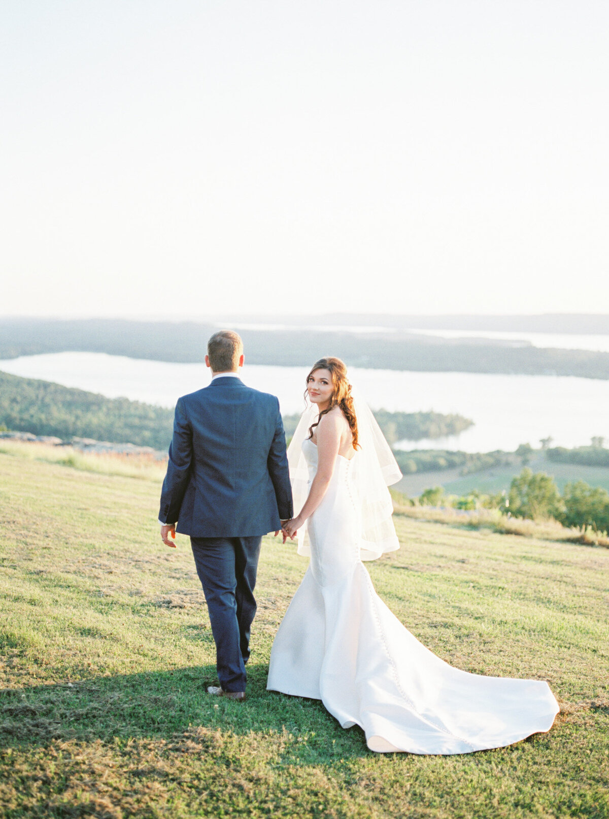 A bride looks back over her shoulder while walking with her groom toward the cliff overlooking a stunning landscape with a lake by Huntsville wedding photographer, Kelsey Dawn Photography