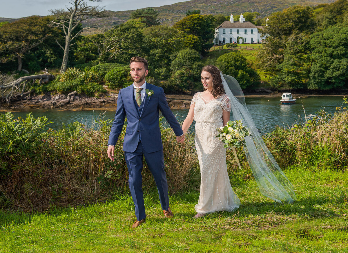 brunette bride wearing vintage, beaded wedding dress, holding a white bouquet and long veil walking with her groom who is wearing a navy suit with Westcove Georgian House in the background