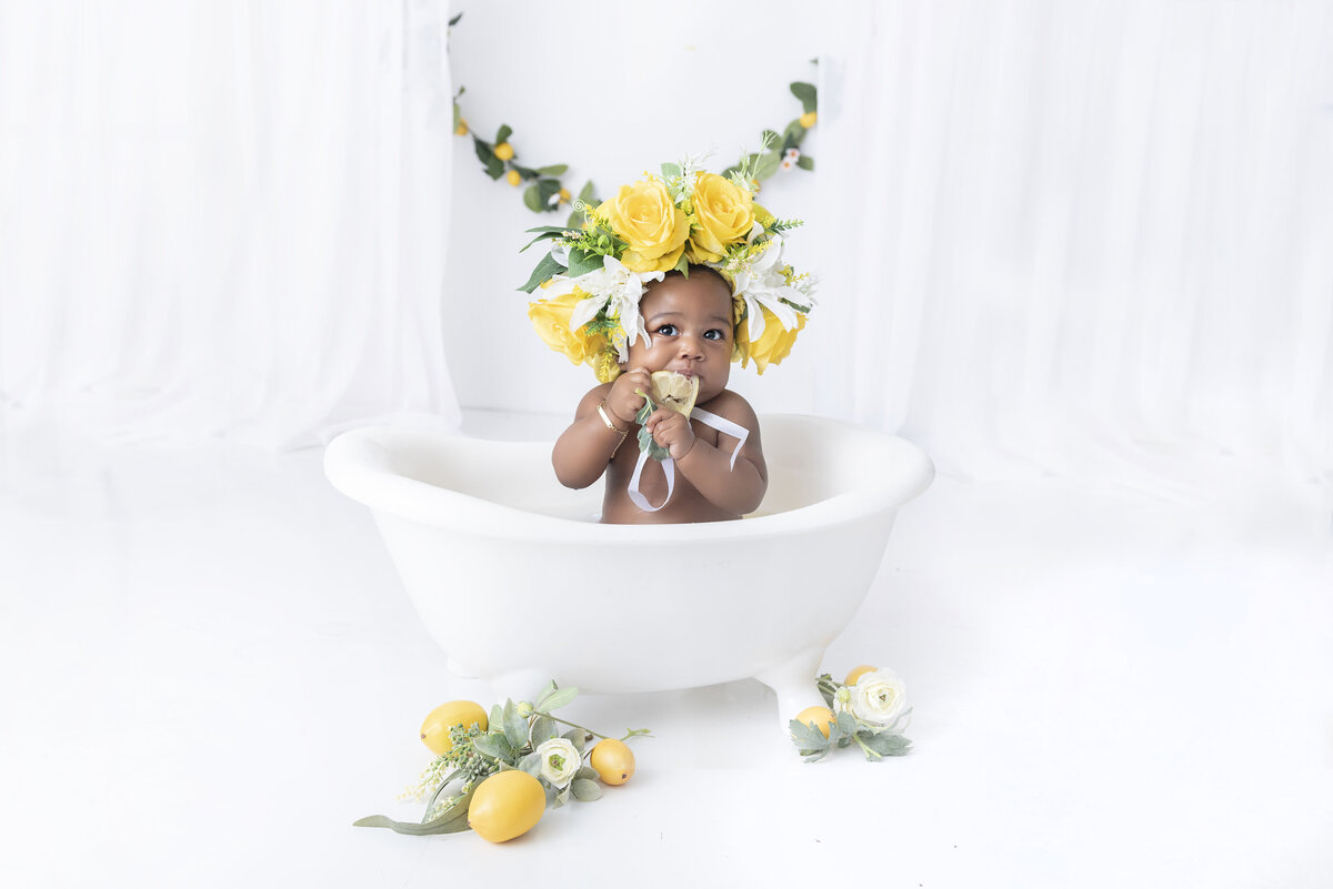 A young toddler girl sits in a bathtub in a studio in a large yellow floral head piece