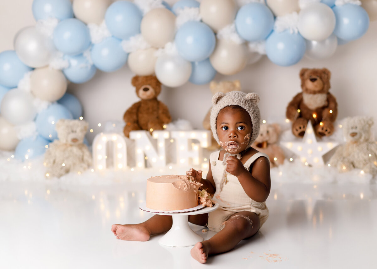 Teddybear picnic themed cake smash at West Palm Beach and Lake Worth newborn photographer. Black baby boy wearing a cream onesie and knit teddy bear hat sitting in front of a beige cake. His face is covered in icing. In the background, there is a blue and white balloon arch and teddy bears.