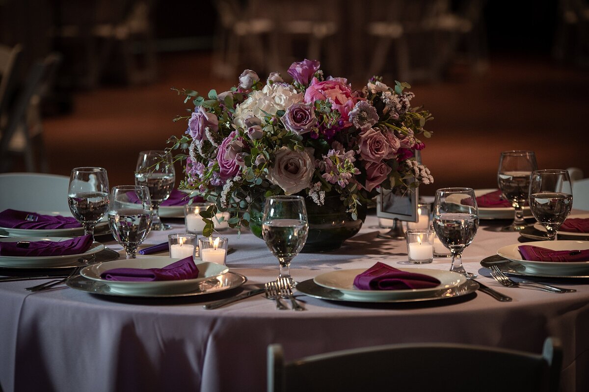 table at Jewish wedding reception with purple linen and large centerpiece with purple roses, pink roses and ivory hydrangea  in a silver bowl. The centerpiece is surrounded by place settings of silver chargers with white plates topped with purple napkins and votive candles at The Liff Center, Nashville