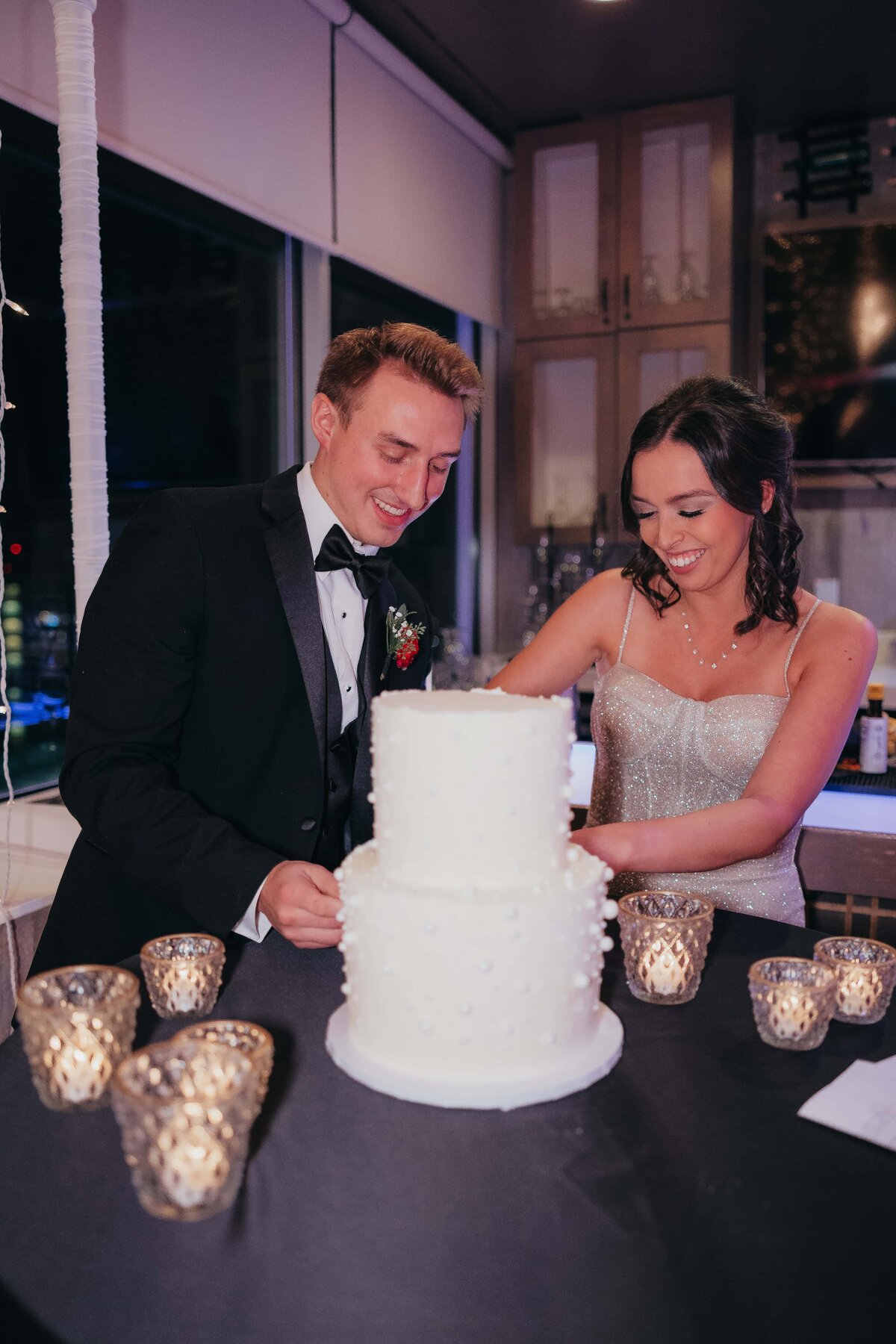 A smiling couple in formal attire cutting a three-tiered wedding cake surrounded by candlelit tables at a reception in a park farm winery.