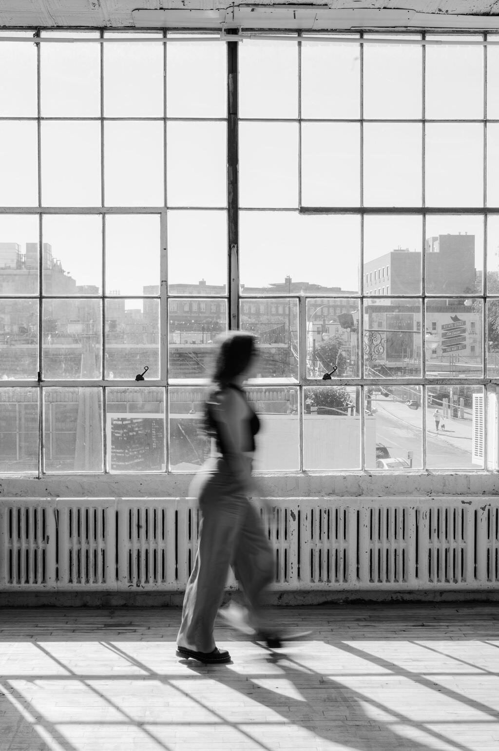 blurred black and white image of a woman walking in an empty room with large windows behind her