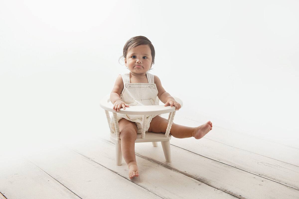 A young toddler sits in a small baby chair in a studio in white overalls