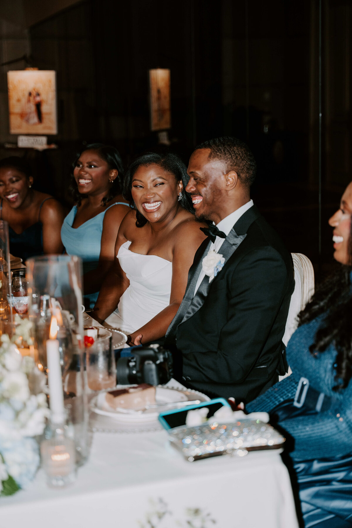 A joyful couple in formal attire is seated at a wedding reception table, surrounded by smiling guests. Elegant decor and candles create a warm ambiance.